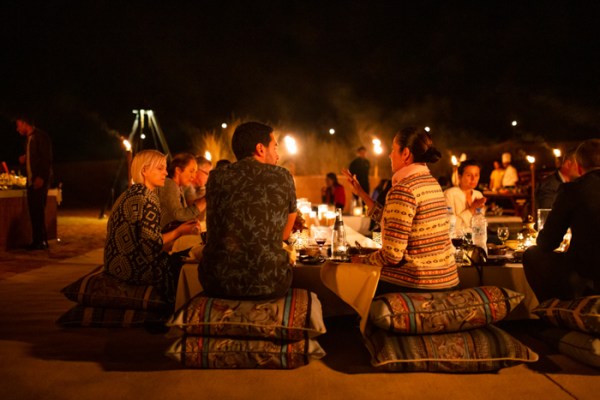 people sitting around a dining table on cushions in the desert at night